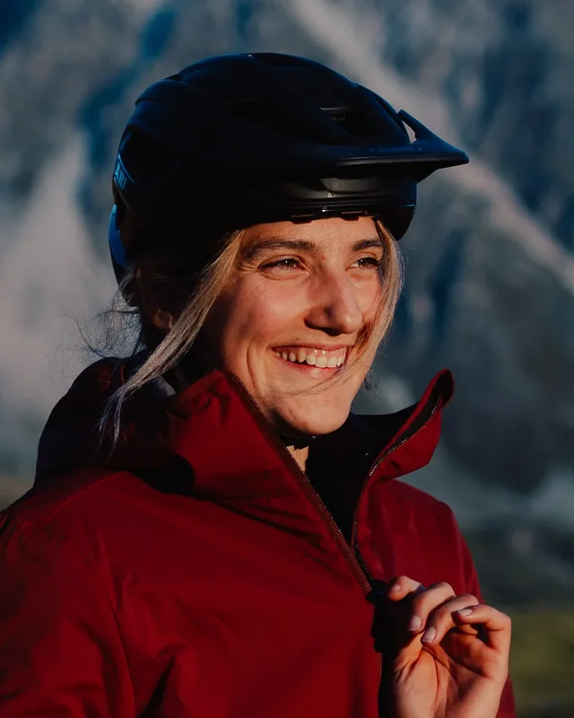 Laura Gut in a red shell and a bike helmet, smiling, with an alpine face behind her
